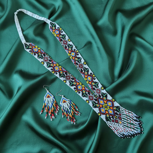 A beaded necklace with matching dangling earrings displayed on a wooden surface. The necklace has a white base with intricate multicolor beadwork in red, yellow, green, blue, and black, and features long fringe at the bottom.