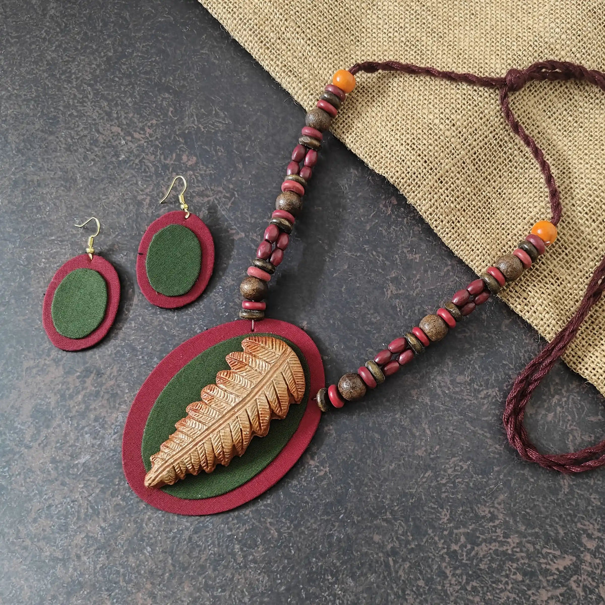 Nirmita maroon fabric necklace set — oval maroon pendant with a green fabric applique and a hand-shaped clay leaf in brown and beige, with oval dangler earrings