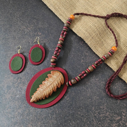 Nirmita maroon fabric necklace set — oval maroon pendant with a green fabric applique and a hand-shaped clay leaf in brown and beige, with oval dangler earrings