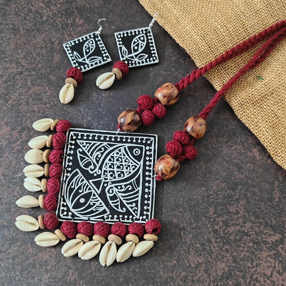 Vyanjana black fabric necklace set — diamond-shaped black pendant with hand-painted lotus and fish motifs, cotton bead and cowrie tassels, white fish earrings