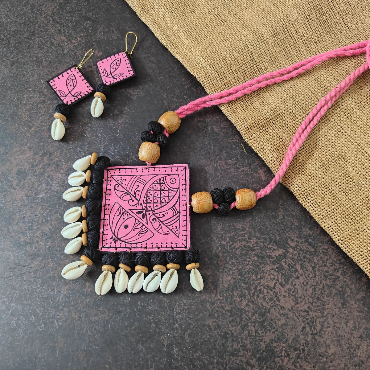 Vyanjana pink fabric necklace set — diamond-shaped pink pendant with lotus and fish hand-painted in black, finished with cotton bead and cowrie tassels